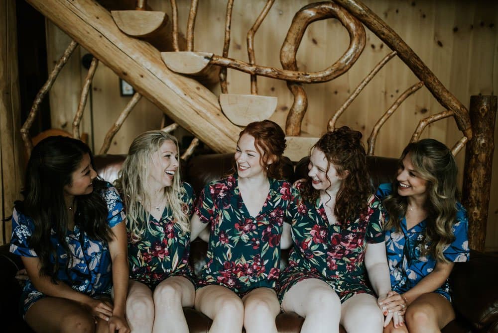 Five women in floral pajamas sit together and smile on a couch with a wooden staircase in the background.