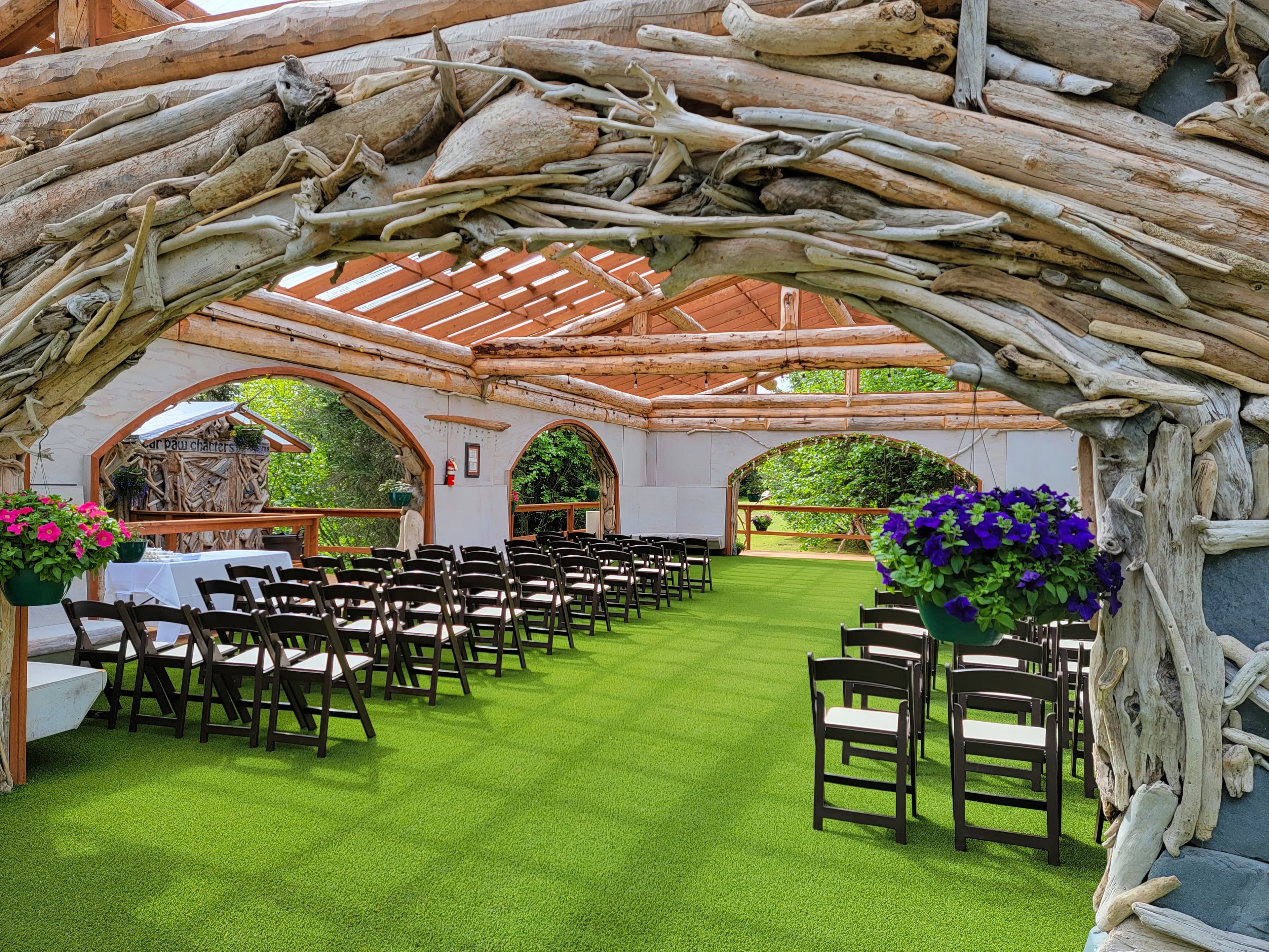 An outdoor venue featuring wooden archways, rows of folding chairs, and vibrant flower planters on green artificial grass.